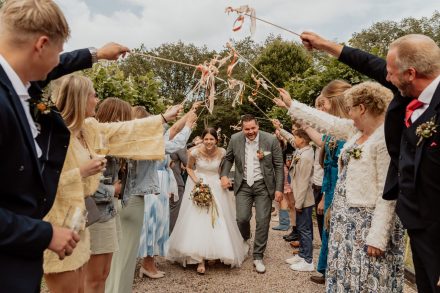 Stralend bruidspaar loopt lachend door een haag van juichende gasten met linten – een prachtig, spontaan trouwmoment.