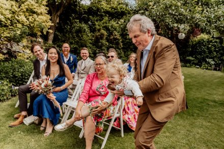 Een vrolijke, speelse momentopname tijdens een bruiloft: een lachende opa tilt zijn kleinkind op, terwijl de gasten genieten van het spontane tafereel.